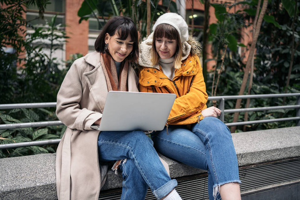 Фото двух белых девушек, использующих ноутбук. Они сидят на каменной скамейке на вокзале.
. - Фото, изображение