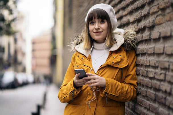 Photo d'une jeune fille caucasienne utilisant son smartphone et écoutant de la musique. Elle s'appuie contre un mur de briques. Elle porte un manteau jaune. Elle sourit.
. - Photo, image
