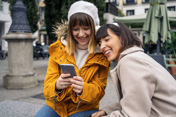 Стокове фото двох кавказьких дівчат, які дивляться на смартфон. Вони сміються. Вони сидять на вулиці
. - Фото, зображення