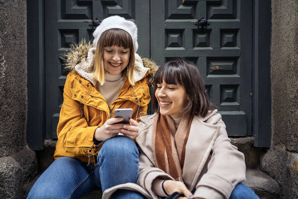 Stock photo of two caucasian girls looking at a smartphone. They are laughing. They are seated on the street. - Fotoğraf, Görsel