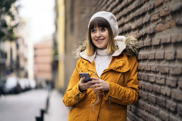 Stockfoto van een jong Kaukasisch meisje dat met haar smartphone naar muziek luistert. Ze leunt tegen een stenen muur. Ze draagt een gele jas. Ze glimlacht.. - Foto, afbeelding