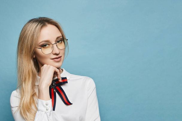Portrait of a young woman with blond hair in white blouse and glasses, isolated at the light blue background with copy space. Smart student girl in white blouse and glasses looking at the camera - Foto, Imagem