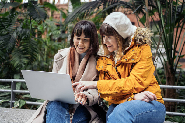 Stock photo of a front view of two caucasian girls using a laptop. They are seated on a stone bench inside a train station. One of them is pointing at the laptop. - Fotoğraf, Görsel