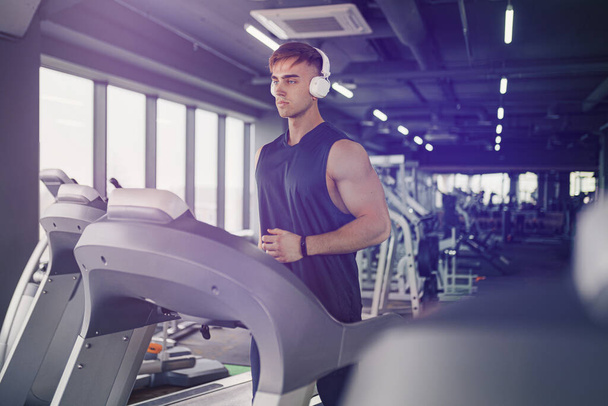 Concentrated fit man listening to music in headphones while running on treadmill in modern gym with panoramic windows. - Zdjęcie, obraz