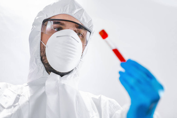 Male scientist wearing mask and protective costume examining thick blood sample in test tube glass in laboratory developing virus research - Photo, Image