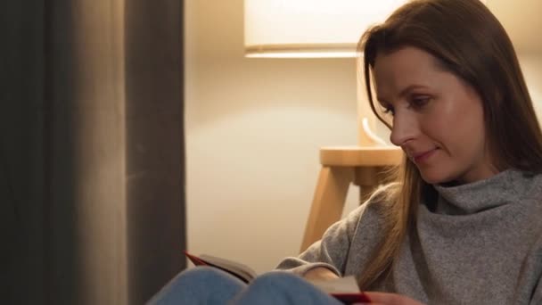Frau liegt auf der Couch in einem gemütlichen Zimmer und liest abends im Schein einer Lampe ein Buch - Filmmaterial, Video