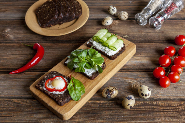 Delicious sandwiches on crusty rye bread with cottage cheese, spinach microgreen, cucumber and baked quail eggs on kitchen board on rustic wooden background - Photo, image