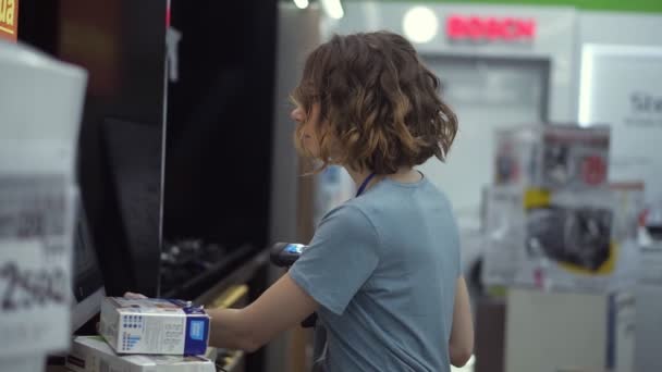 Curly haired concentrated woman seller-consultant in household department carry and scan by barcode stationery goods in the boxes, standing in front the screen. Slow motion. Side view - Footage, Video