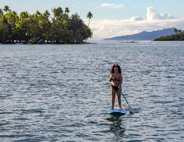 Active woman with curly hair stand up paddleboard on the sunset near the island of Bora Bora in the Pacific Ocean, French Polynesia - Фото, изображение