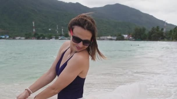 Ein Mädchen hält an einem tropischen Strand einen Pareo in den Händen. Eine junge Frau im Badeanzug mit Seidentuch spaziert an der Küste entlang. - Filmmaterial, Video