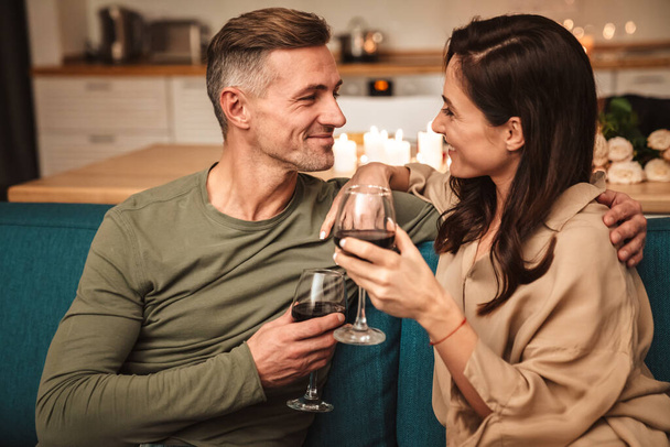 Image of happy passionate couple drinking red wine from glasses while having romantic candlelight dinner at home - Photo, image