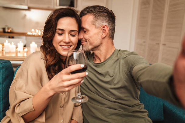 Image of happy caucasian couple taking selfie photo while having romantic candlelight dinner at home - Foto, Bild
