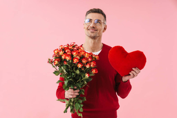 Portrait of handsome caucasian man wearing eyeglasses holding flowers and toy heart isolated over pink background - Foto, afbeelding