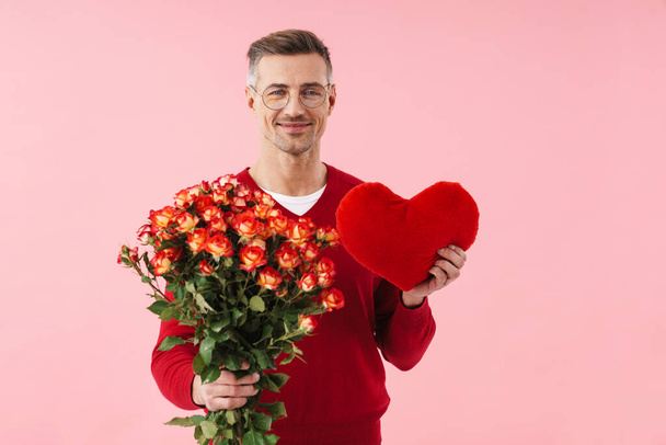 Portrait of handsome caucasian man wearing eyeglasses holding flowers and toy heart isolated over pink background - Foto, immagini
