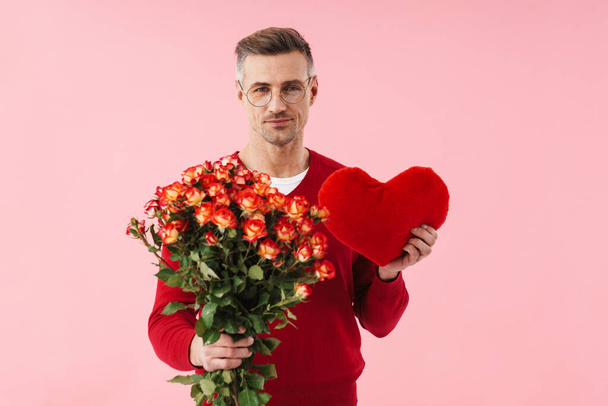 Portrait of handsome caucasian man wearing eyeglasses holding flowers and toy heart isolated over pink background - Fotoğraf, Görsel