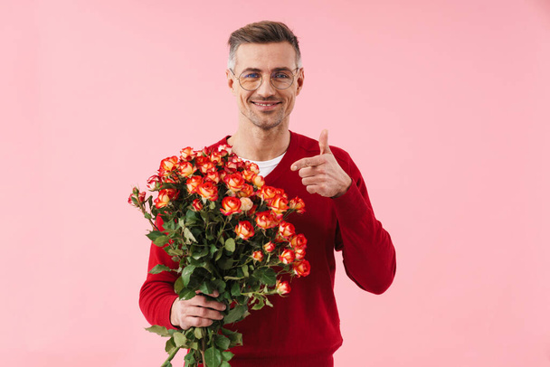 Portrait of handsome caucasian man wearing eyeglasses pointing finger at flowers in his hand isolated over pink background - Фото, изображение