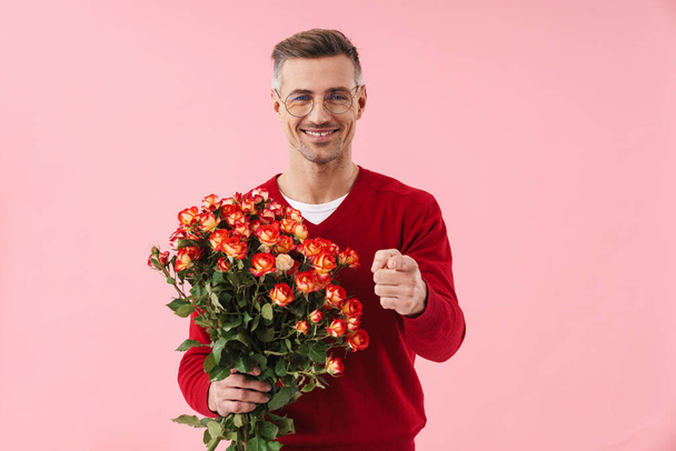 Portrait of handsome caucasian man wearing eyeglasses holding flowers and pointing finger at camera isolated over pink background - Фото, изображение