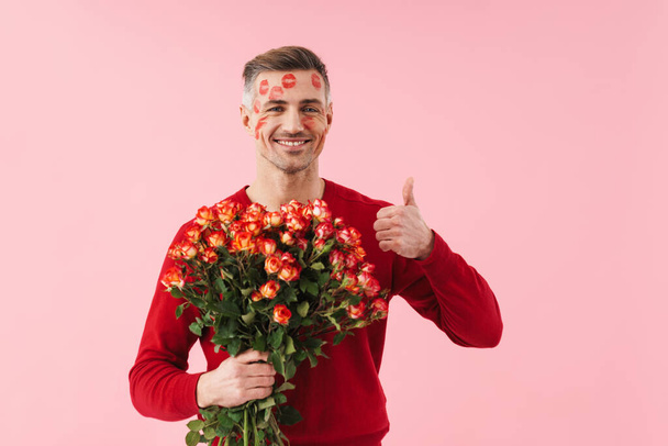 Portrait of handsome caucasian man with kiss marks at his face holding flowers on valentines day isolated over pink background - Photo, image