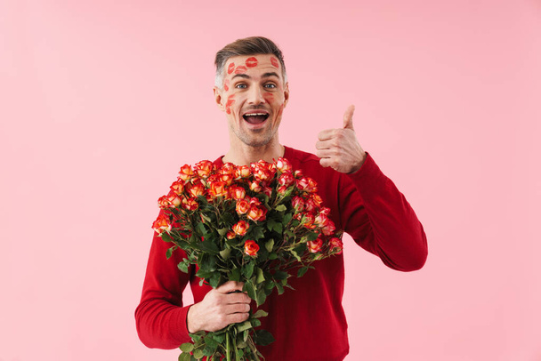 Portrait of handsome caucasian man with kiss marks at his face holding flowers on valentines day isolated over pink background - Fotoğraf, Görsel