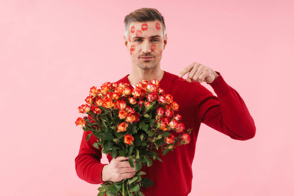 Portrait of handsome caucasian man with kiss marks at his face holding flowers on valentines day isolated over pink background - Foto, afbeelding