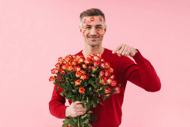 Portrait of handsome caucasian man with kiss marks at his face holding flowers on valentines day isolated over pink background - Photo, image