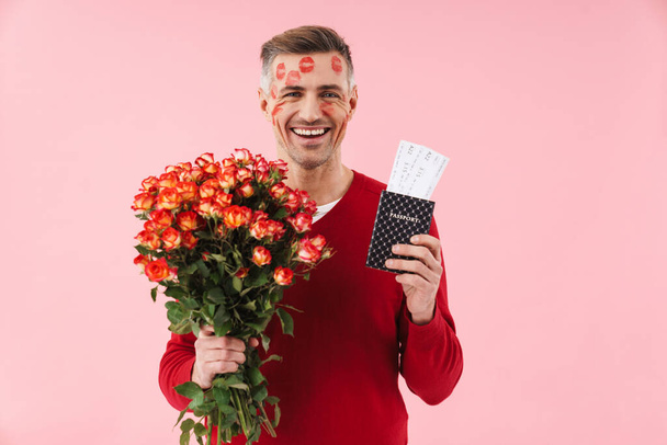 Portrait of handsome caucasian man with kiss marks at his face holding flowers and travel tickets isolated over pink background - Foto, Bild