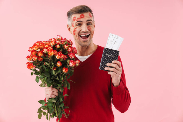 Portrait of handsome caucasian man with kiss marks at his face holding flowers and travel tickets isolated over pink background - Φωτογραφία, εικόνα