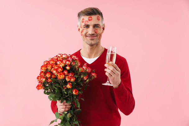 Portrait of handsome caucasian man with kiss marks at his face holding flowers and champagne glass isolated over pink background - Photo, Image