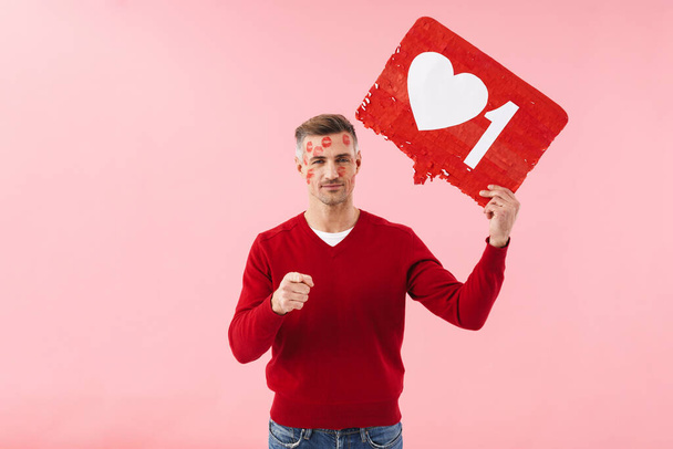 Portrait of handsome caucasian man holding heart like symbol on placard and pointing finger at camera isolated over pink background - Foto, afbeelding