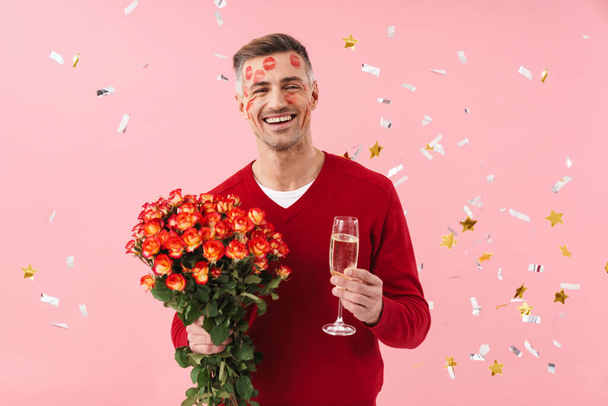 Image of a positive happy mature man with lipstick on face isolated over pink wall background in red sweater holding flowers and champagne over confetti. - Photo, Image