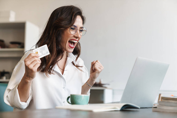 Schöne junge brünette Frau mit Kreditkarte, während sie drinnen am Cafétisch mit Laptop-Computer sitzt und Erfolg feiert - Foto, Bild