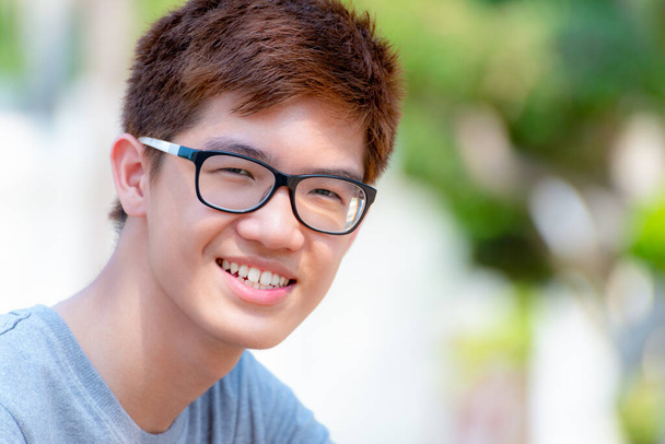 Closeup face Asian handsome teenage boy wearing glasses, Portrait cheerful hipster young man are smiling with a happy face at park on green nature background in Thailand - Photo, Image