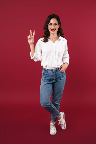 Photo of a smiling young amazing woman posing isolated over red wall background showing peace gesture. - Fotó, kép