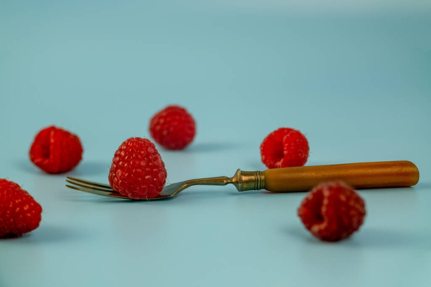 raspberries scattered on a blue background, one lying on a beautiful, antique fork - Photo, Image