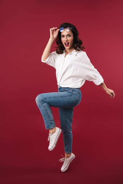 Photo of a pretty beautiful young woman jumping isolated over red wall background. - Photo, Image