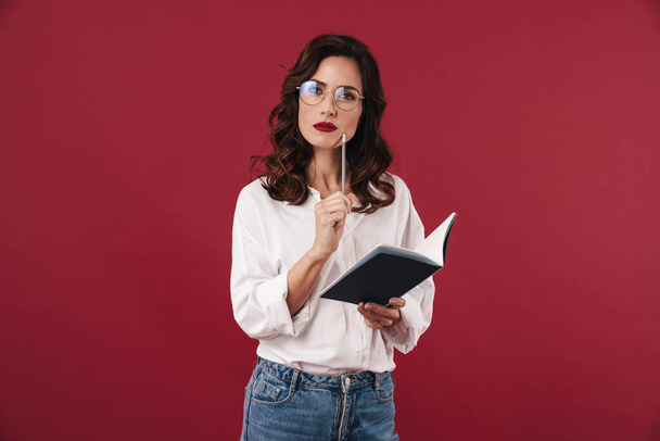 Image of serious thinking young woman in glasses isolated over red wall background writing notes in notebook. - Фото, изображение