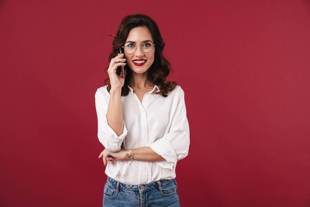 Image of optimistic smiling young woman in glasses isolated over red wall background talking by mobile phone. - Foto, afbeelding