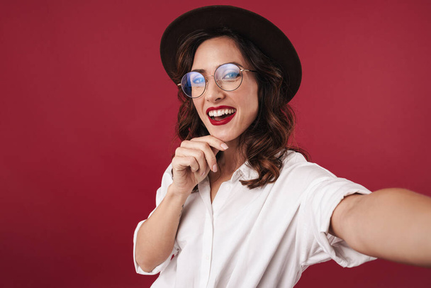 Picture of cheery optimistic happy young woman in glasses isolated over red wall background take a selfie by camera. - Foto, afbeelding
