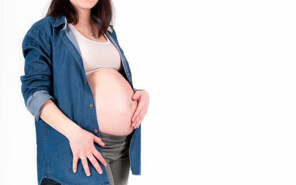 Teilaufnahme einer schwangeren Frau in blauem Jeanshemd und oben stehendem und anrührendem Bauch isoliert auf weißem Grund - Foto, Bild