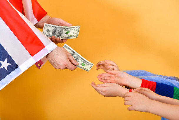 Mano de un hombre bajo la bandera americana manos dinero en manos de niños, concepto de dependencia de los EE.UU.
. - Foto, Imagen