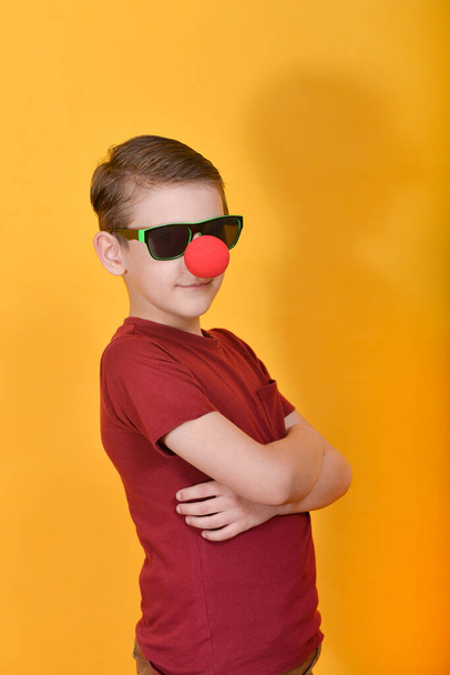 Een serieuze jongen met een clown neus staat op een gele achtergrond met zijn handen gevouwen in de buurt van zijn borst. - Foto, afbeelding