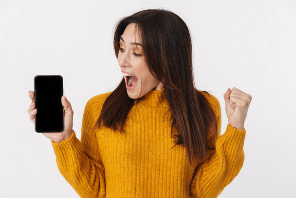 Wizerunek pięknej brunetki dorosłej kobiety noszącej sweter krzycząc i trzymając telefon komórkowy na białym tle - Zdjęcie, obraz