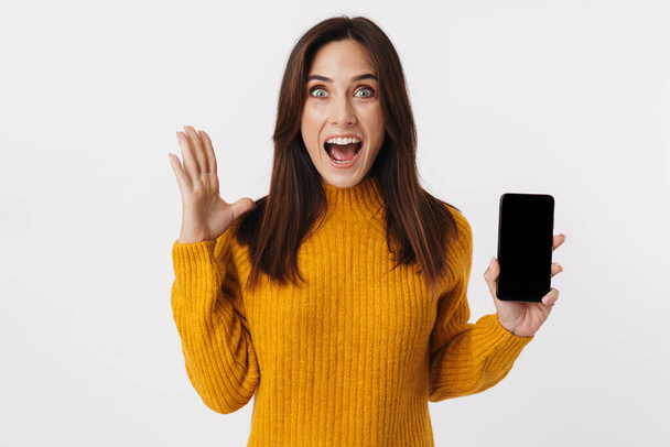 Image of beautiful brunette adult woman wearing sweater screaming and holding cellphone isolated over white background - Fotó, kép