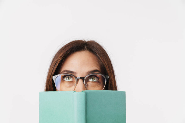 Image of beautiful brunette adult woman wearing eyeglasses looking upward and covering her face with book isolated over white background - Valokuva, kuva