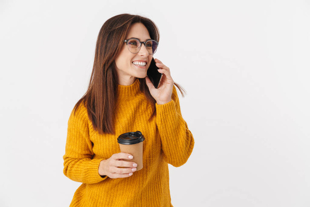 Image of beautiful brunette adult woman wearing eyeglasses holding coffee cup and talking on cellphone isolated over white background - 写真・画像