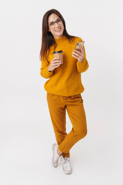 Full length image of beautiful brunette adult woman wearing eyeglasses holding coffee cup and cellphone isolated over white background - Фото, изображение