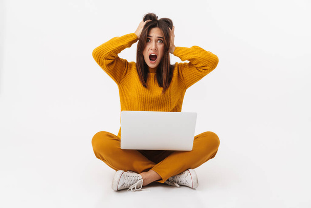 Bild der schönen brünetten erwachsenen Frau ergreift Kopf und hält Laptop-Computer, während sie mit überkreuzten Beinen isoliert auf weißem Hintergrund sitzt - Foto, Bild