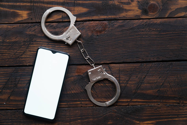 smartphone with blank screen for your text or logo with handcuffs rests on a wooden background - Foto, Imagem
