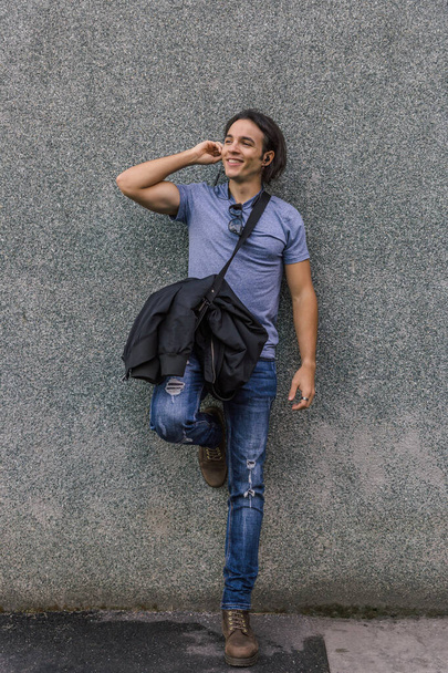 Kaukasischer gutaussehender Mann in blauem T-Shirt und Jeans lächelt und hört Musik, die sich im Frühling an eine graue Wand lehnt - Foto, Bild