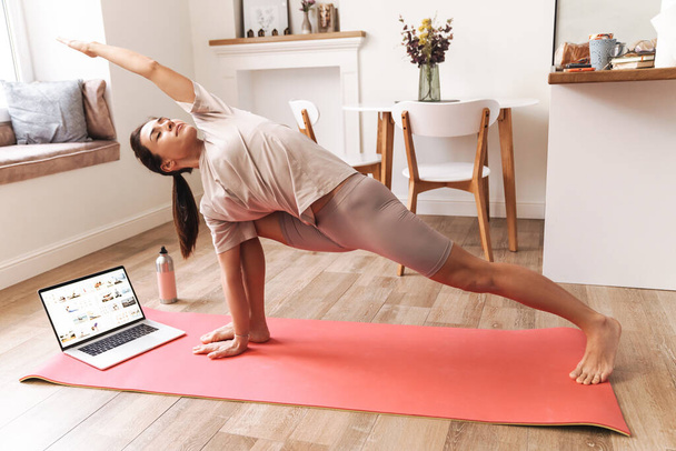 Image of a concentrated young beautiful brunette fitness woman make stretching exercises indoors at home near laptop computer. - Foto, Bild